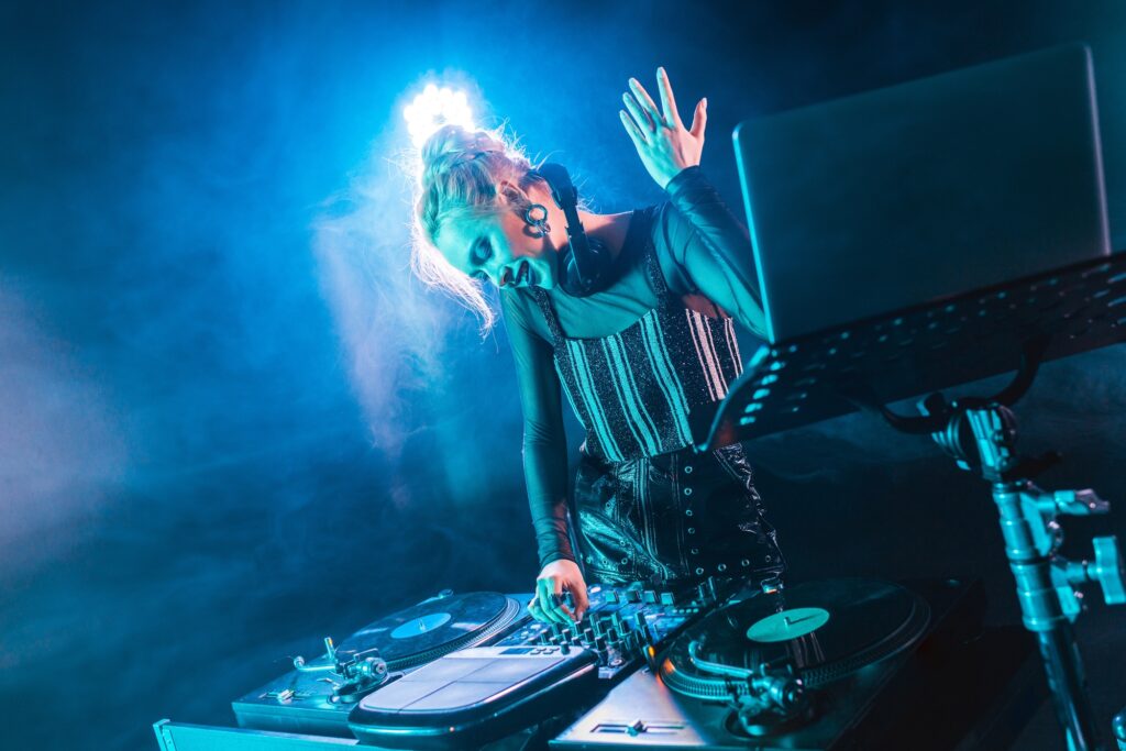 smiling blonde dj girl in headphones standing near dj mixer and gesturing in nightclub with smoke